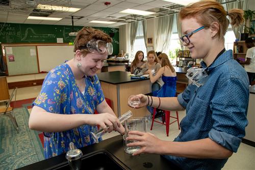 Two high school students pouring clear liquid in science beakers 